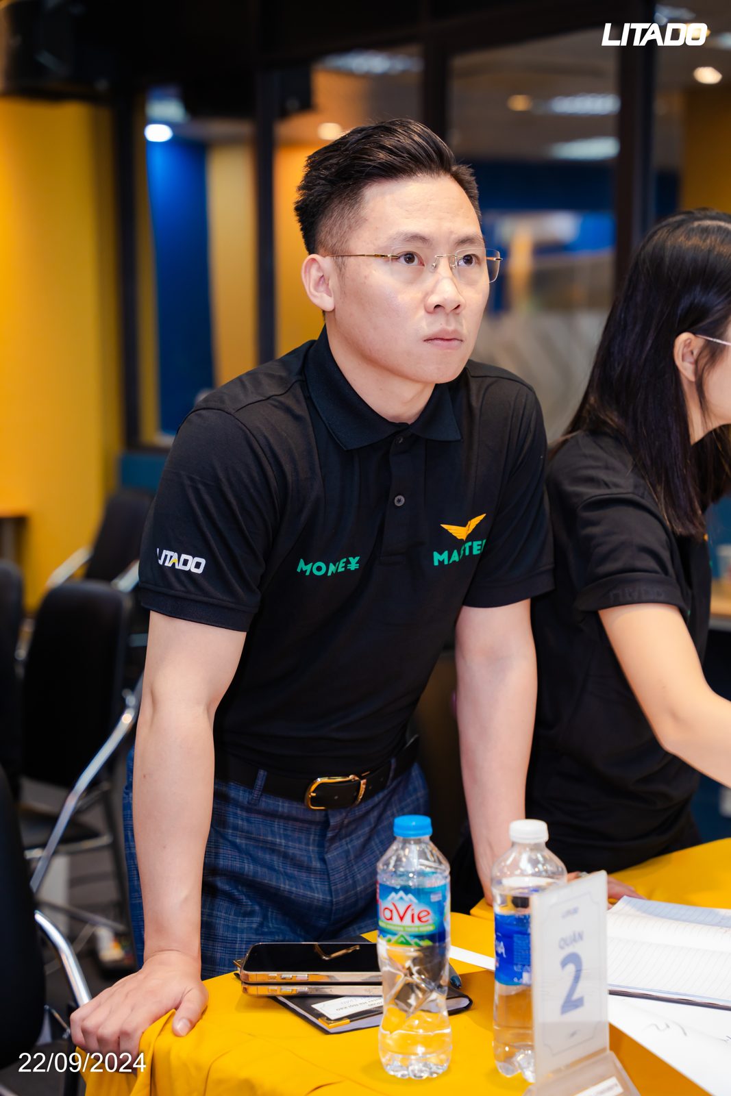 Lê Anh Tuấn mặc áo polo đen đứng bên bàn học trong một lớp học đầu tư