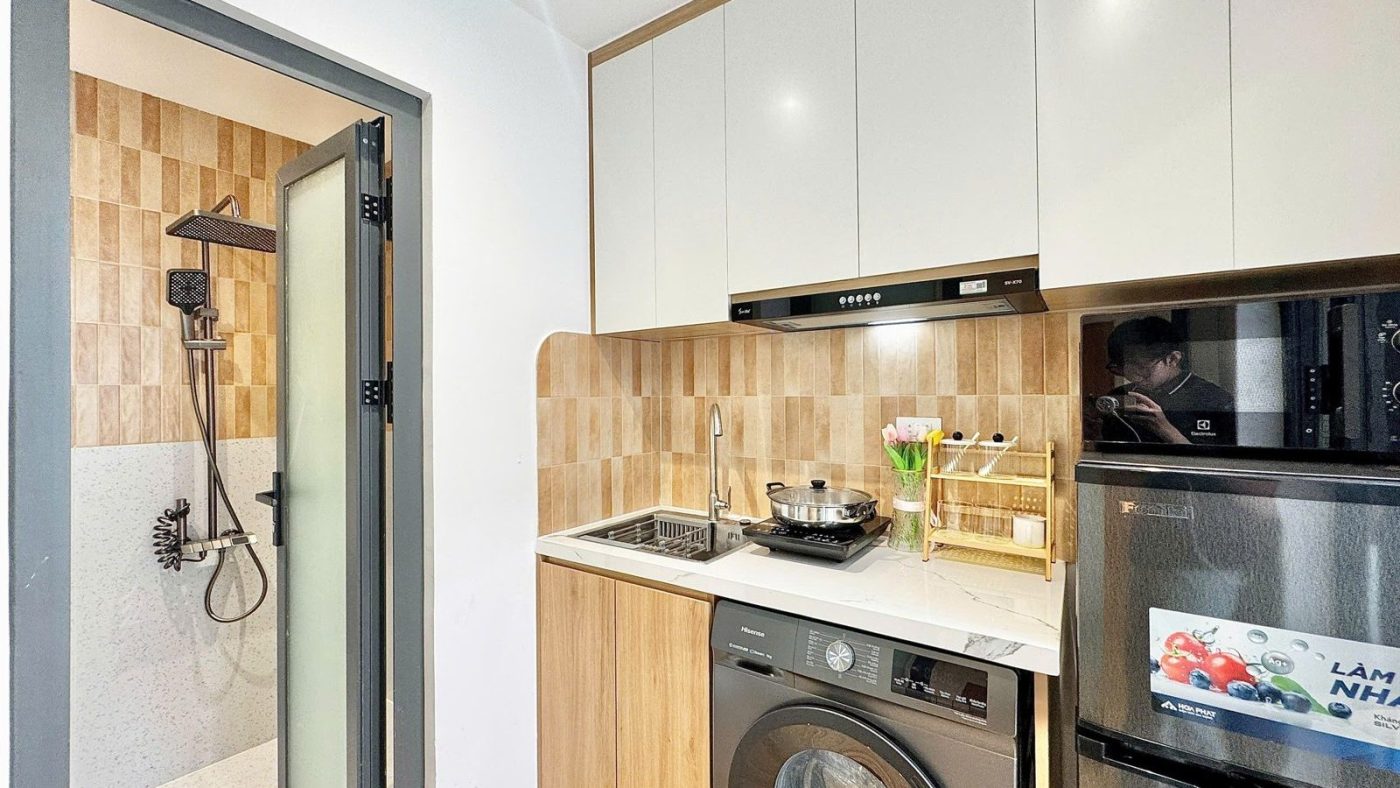 Góc bếp tủ trắng, bàn bếp, máy giặt và tủ lạnh nhỏ cạnh cửa phòng tắm trong một phòng cho thuê, ảnh bìa bài viết về bán căn hộ dịch vụ Tân Bình