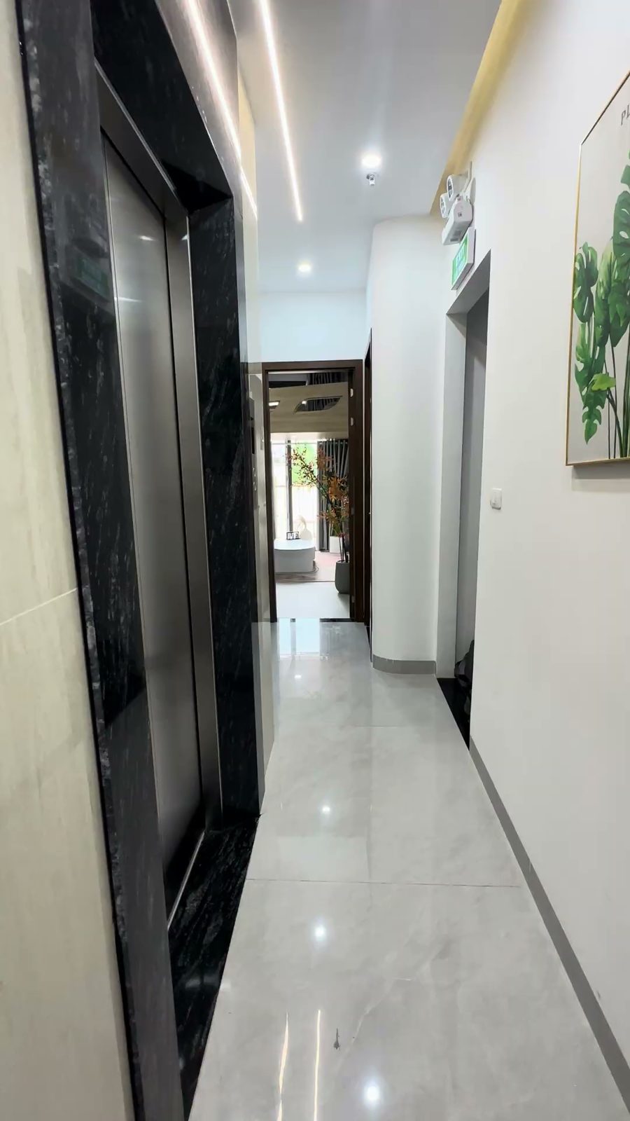 Hành lang lát đá sáng màu có cửa thang máy đen và tranh lá cây trên tường