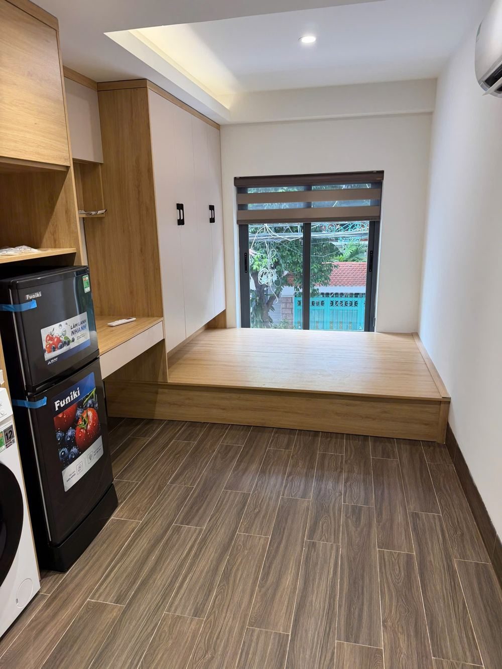 Phòng cho thuê lát sàn gỗ có bục, bếp, tủ lạnh và máy giặt, cửa sổ nhìn ra cây xanh