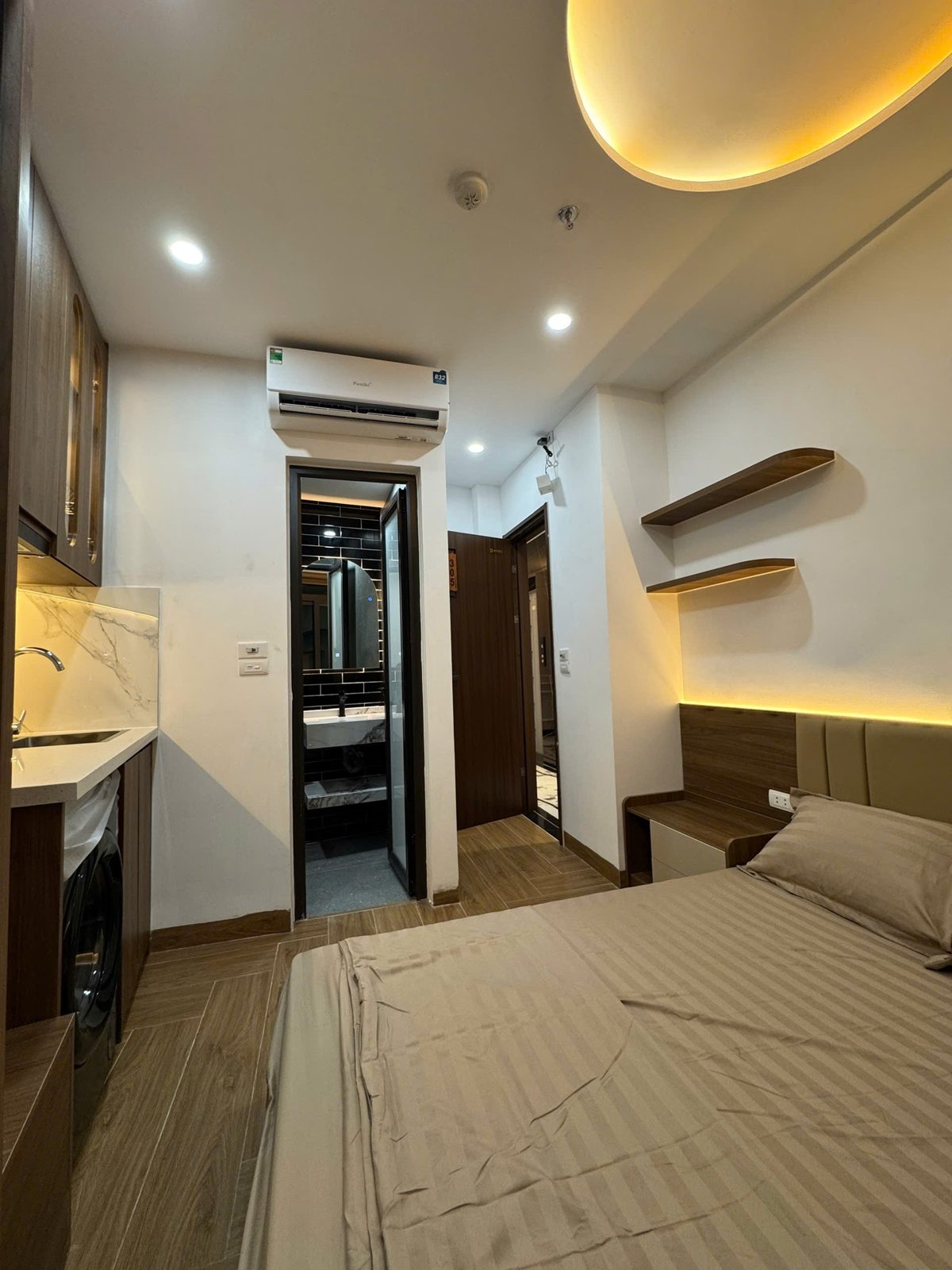 Phòng căn hộ dịch vụ có giường, kệ gỗ treo tường, điều hòa và lối vào nhà tắm
