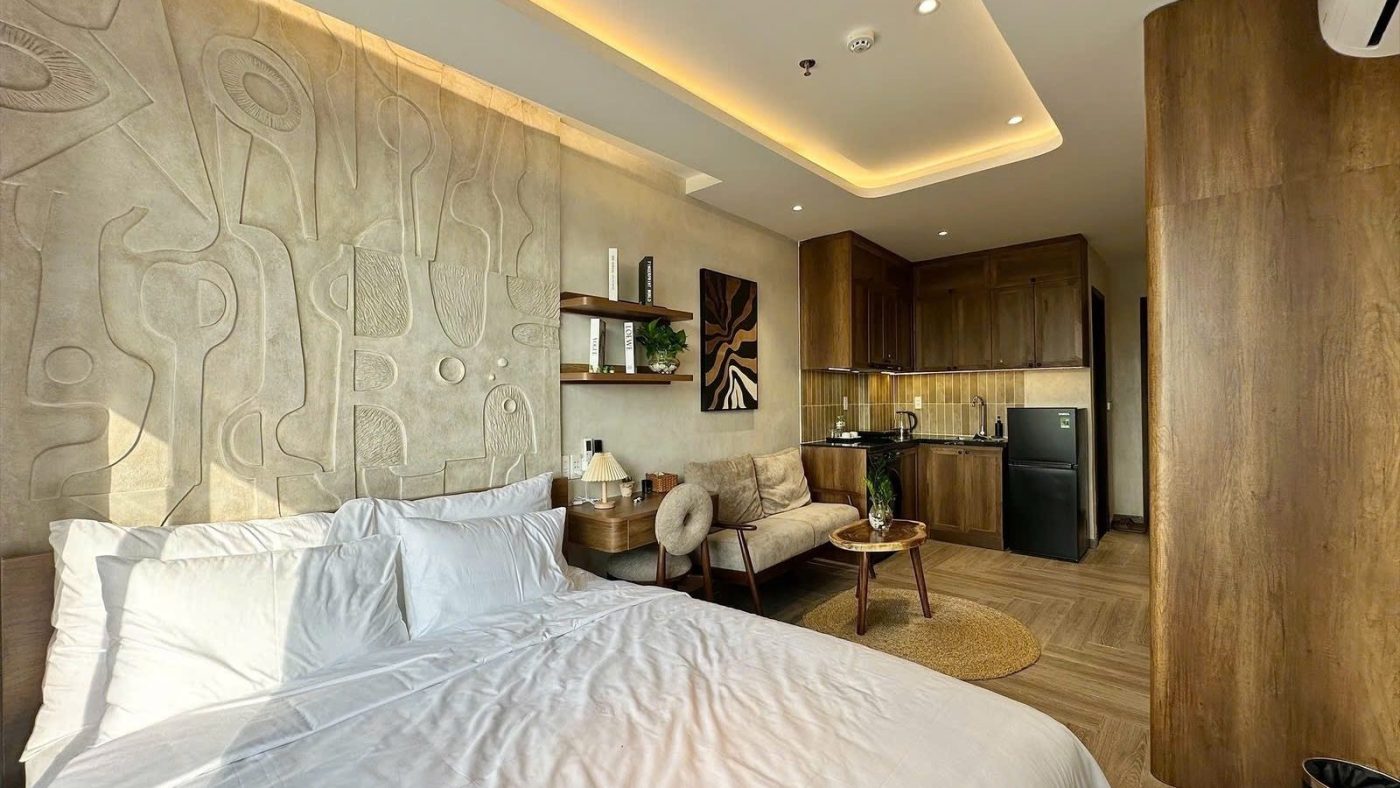 Giường trải ga trắng trước mảng tường phù điêu, phía sau là sofa, kệ gỗ và góc bếp