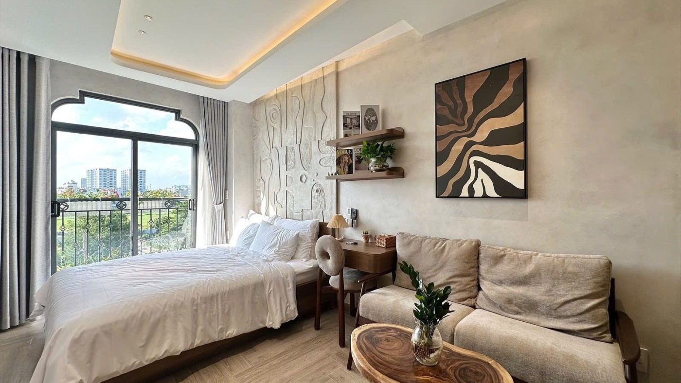 Phòng cho thuê có giường, sofa vải, bàn trà gỗ, tranh trừu tượng và cửa sổ vòm nhìn ra thành phố