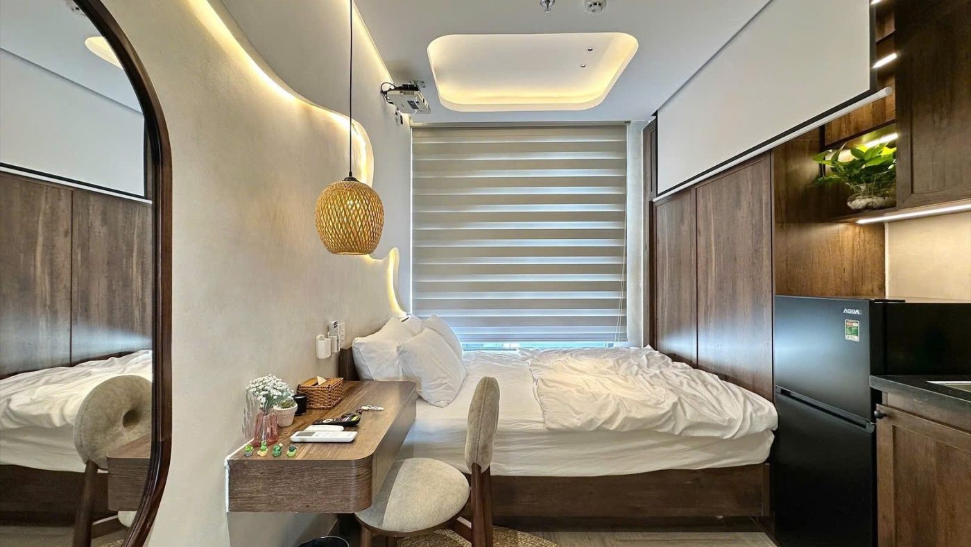 Phòng cho thuê nội thất gỗ có đèn thả mây, gương bầu dục, bàn làm việc và giường cạnh cửa sổ rèm cuốn