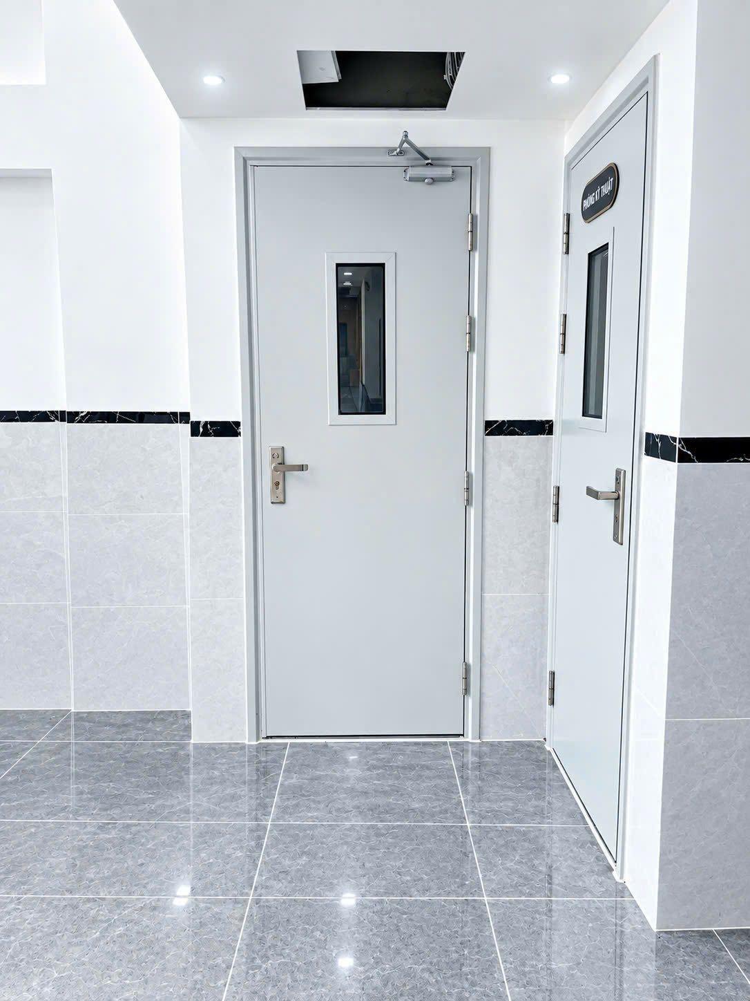 Hai cánh cửa thép sơn xám có ô kính và tay co tự đóng ở tầng trệt một tòa nhà