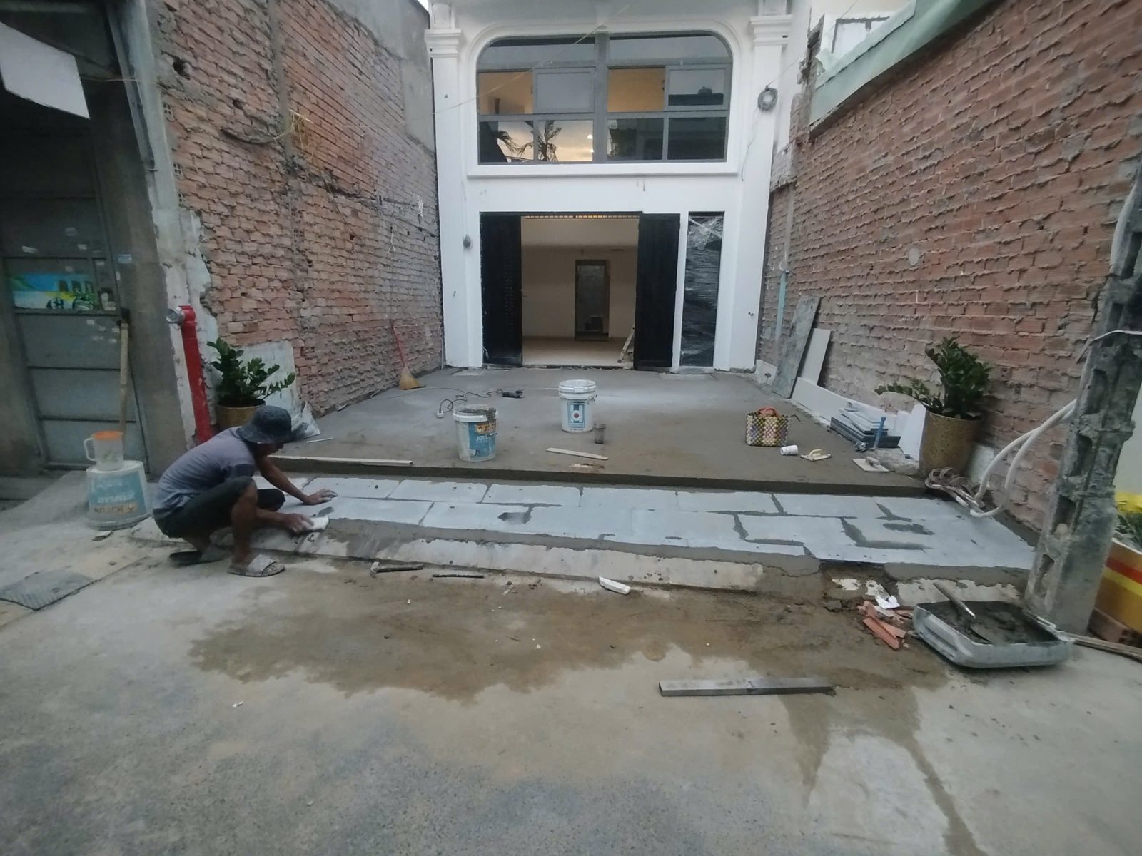 Khoảng sân trước một tòa nhà đang hoàn thiện, thợ đang lát nền, hai bên là tường gạch của nhà hàng xóm