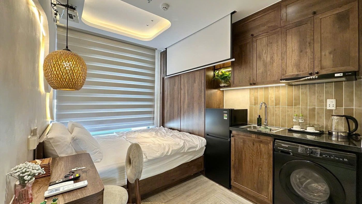 Phòng cho thuê có đèn thả mây, giường cạnh cửa sổ, bếp gỗ kèm tủ lạnh và máy giặt