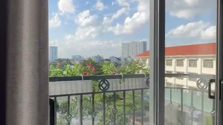 Nhìn qua cửa kính ban công ra hàng cây và một ngôi trường mái đỏ dưới trời xanh