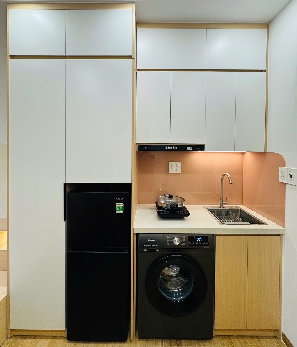 Góc bếp phòng cho thuê có tủ lạnh đen, máy giặt, bếp và tủ bếp trắng