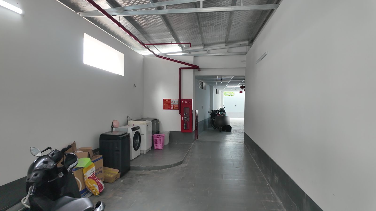Khu để xe tầng trệt có máy giặt chung, tủ chữa cháy và đường ống đỏ chạy dọc trần