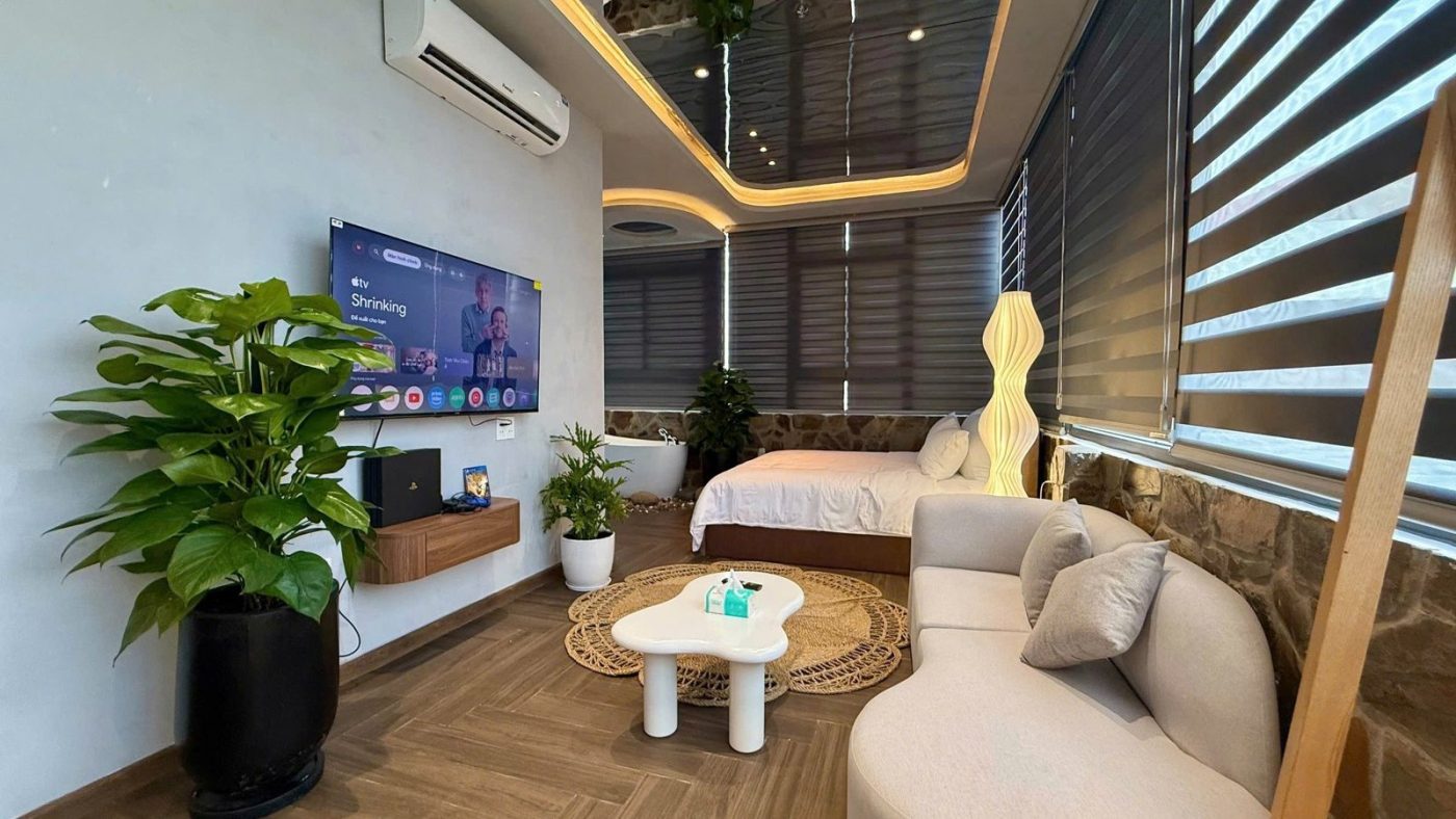 Phòng căn hộ dịch vụ rộng có sofa trắng, tivi treo tường, cây xanh và rèm cuốn