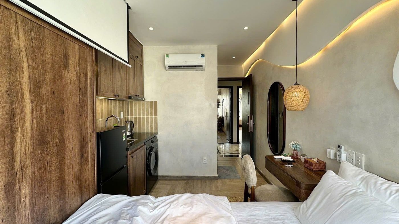 Phòng cho thuê nhìn từ giường thấy tường ốp gỗ, góc bếp, gương bầu dục, đèn thả mây và điều hòa, ảnh bìa bài viết về nhà cho thuê lợi nhuận cao