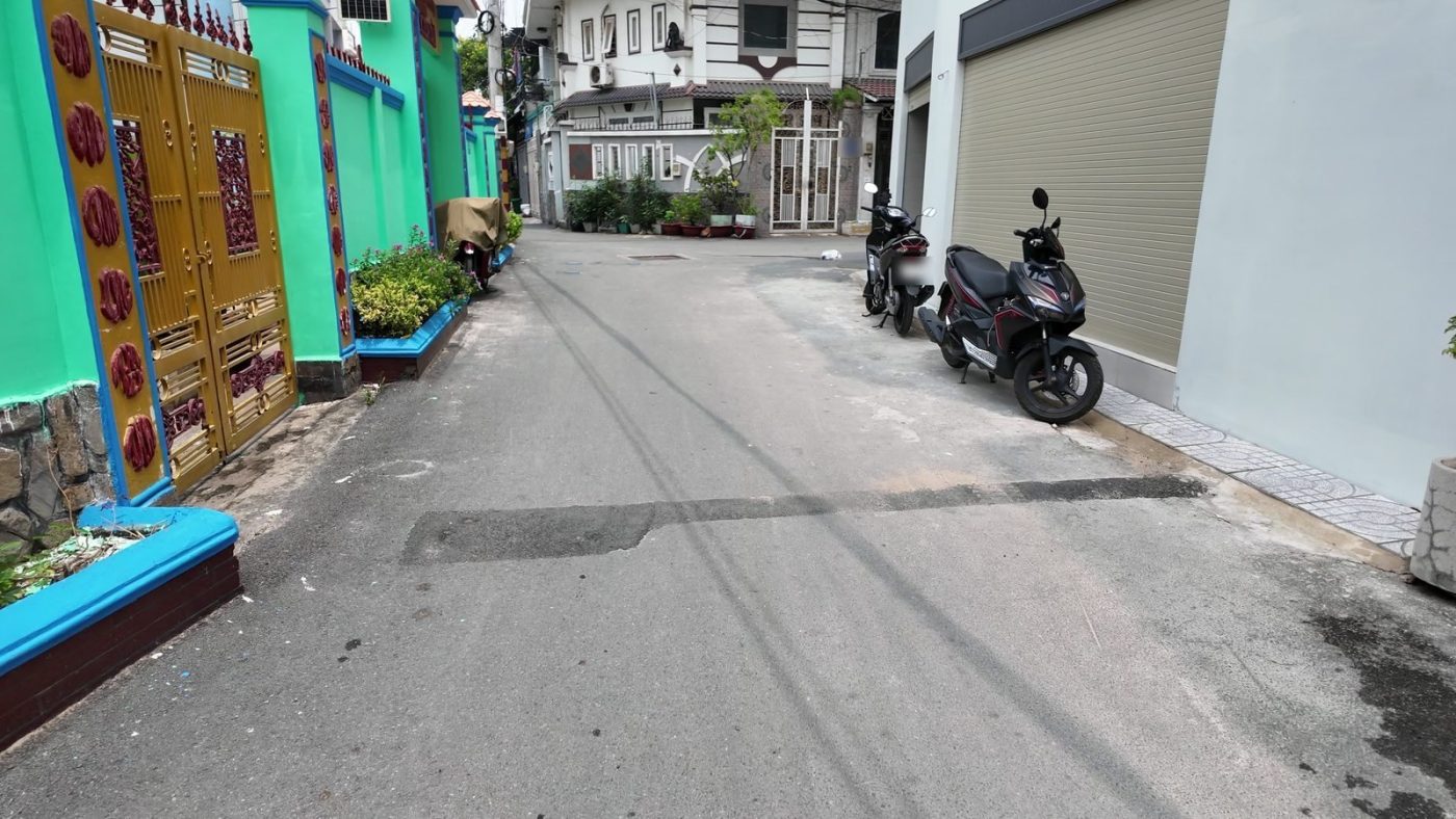 Con hẻm rộng dẫn ra trục chính ở Gò Vấp, một bên là tường rào xanh, có xe máy dựng ven đường