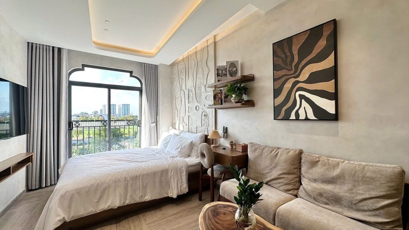 Phòng cho thuê nội thất gỗ có giường, sofa, bàn trà, tranh trừu tượng và cửa vòm nhìn ra ban công, ảnh bìa bài viết về nhà hẻm hay nhà mặt tiền