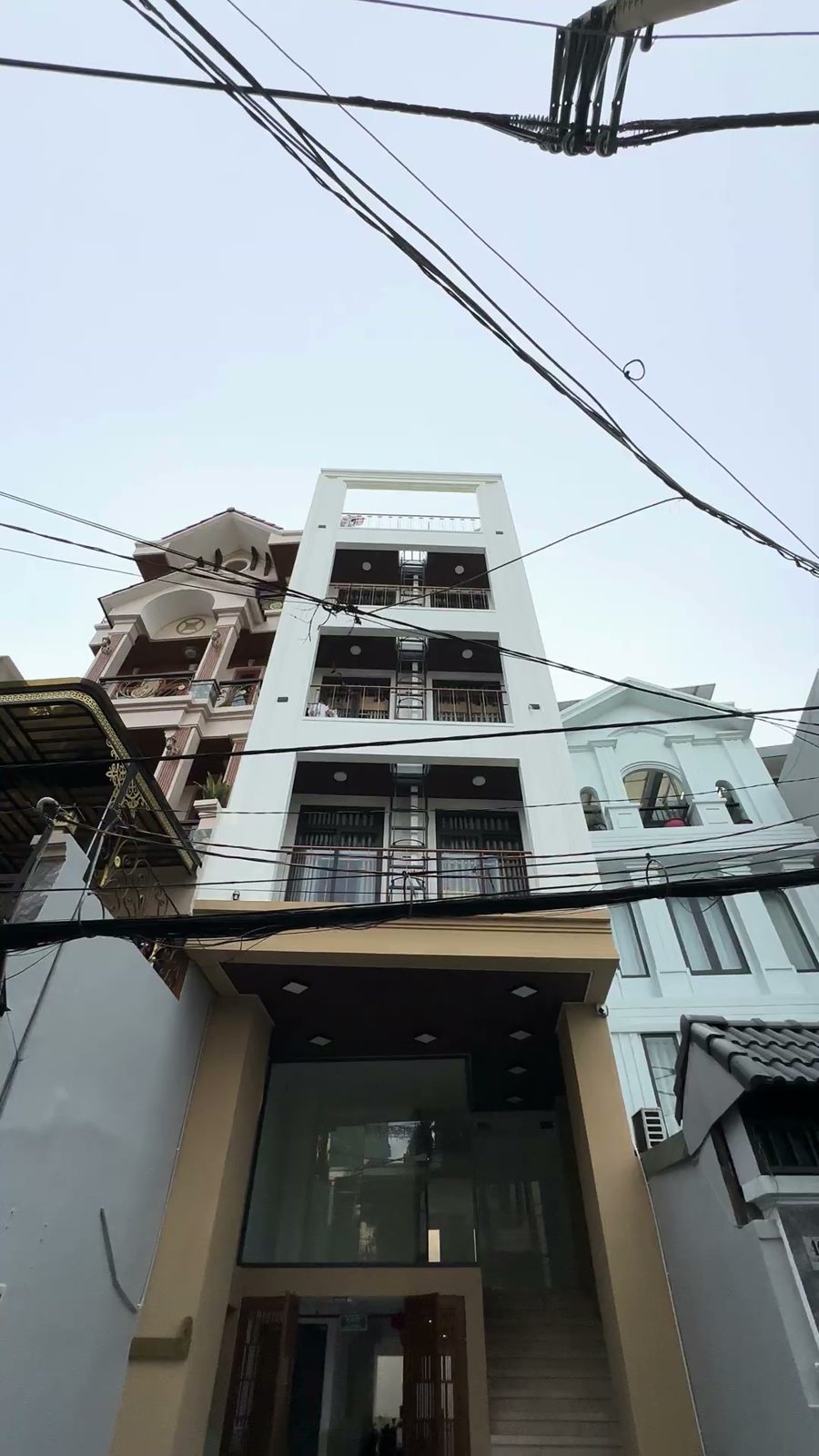 Tòa nhà nhiều tầng sơn trắng nằm giữa các nhà dân trong hẻm, dây điện giăng phía trước