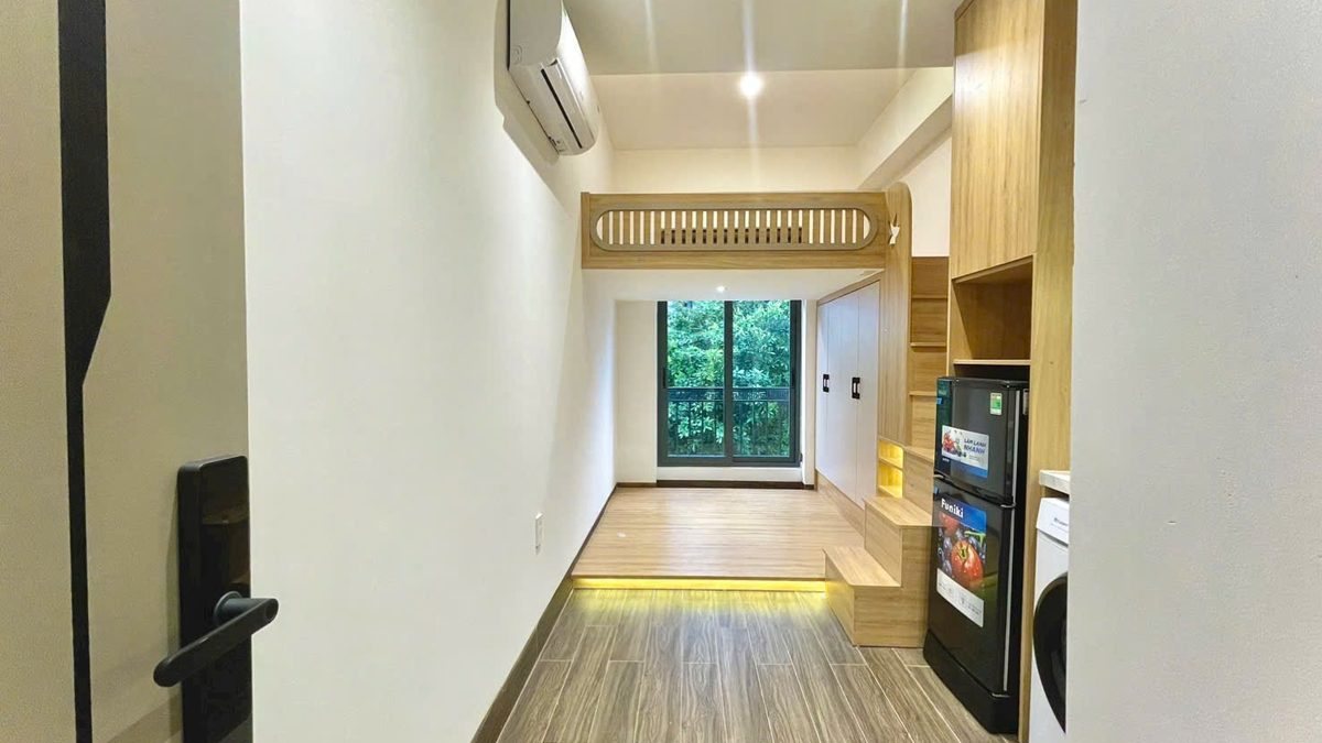 Phòng cho thuê dài có sàn gỗ, điều hòa, gác gỗ, tủ lạnh và cửa kính cuối phòng nhìn ra cây xanh