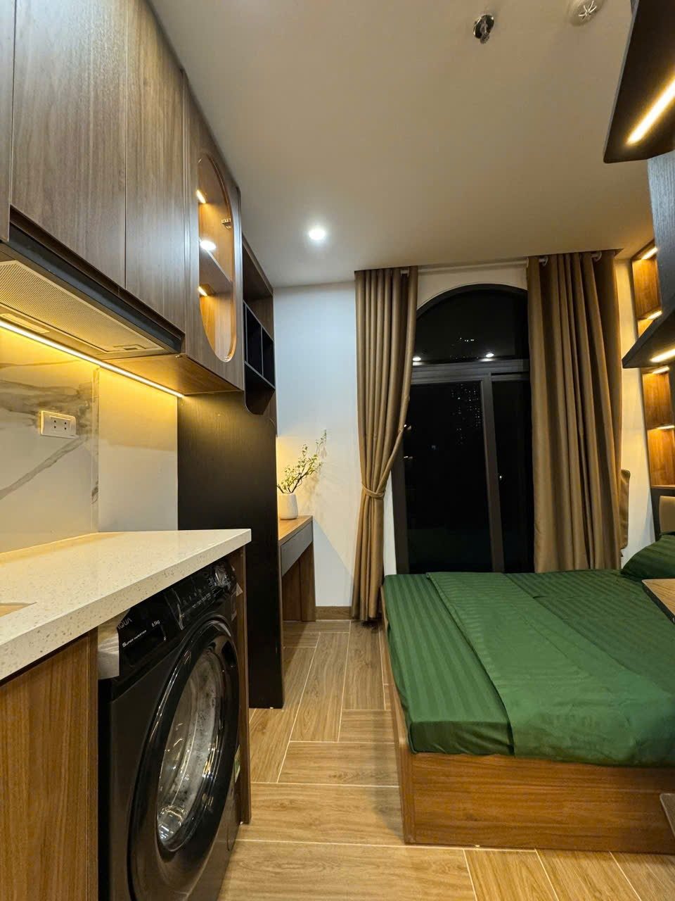 Phòng căn hộ dịch vụ có giường trải ga xanh, bếp, máy giặt và rèm cửa vàng