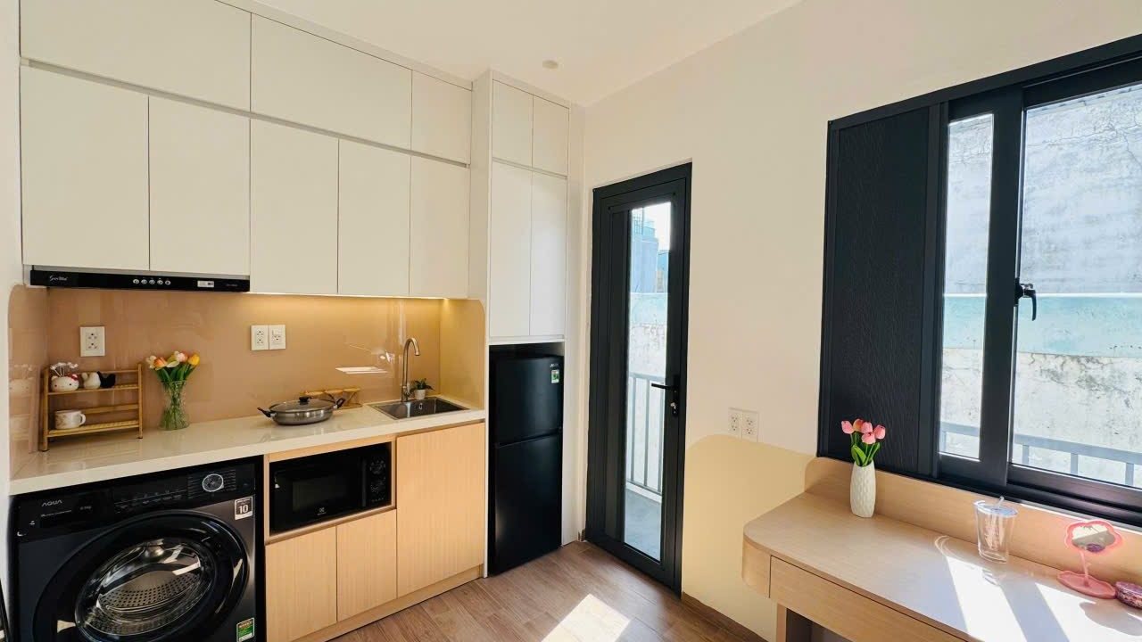 Góc bếp có máy giặt, tủ lạnh và bàn làm việc cạnh cửa sổ trong một phòng cho thuê, ảnh bìa bài viết về tự vận hành căn hộ dịch vụ