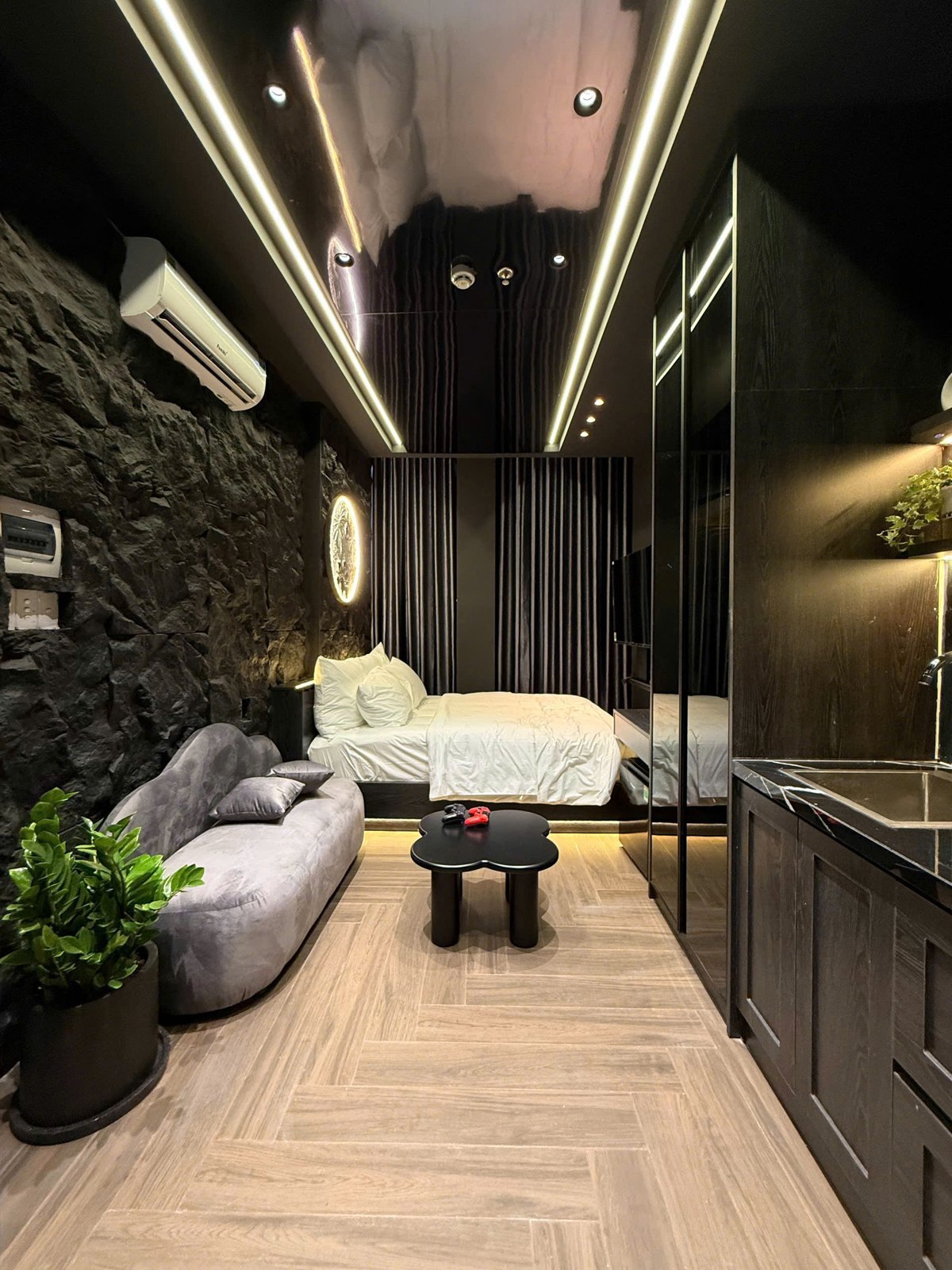 Phòng cho thuê có mảng tường đá tối màu, đèn tròn, sofa xám và giường ở cuối phòng