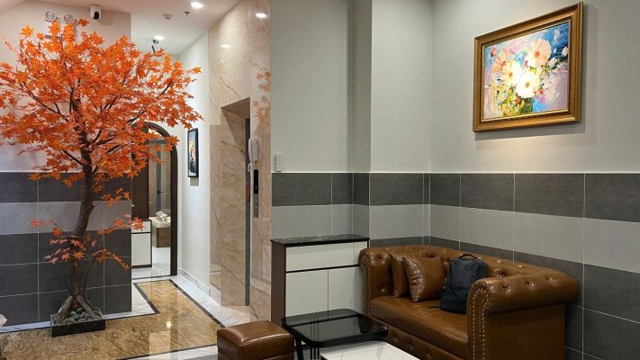 Sảnh nhỏ có cây phong lá cam, ghế sofa da, tranh hoa và cửa thang máy phía sau, ảnh bìa bài viết về vợ chồng vay mua nhà